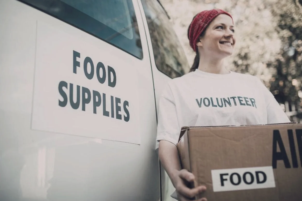 A smiling volunteer carrying a box of food beside a food supplies van