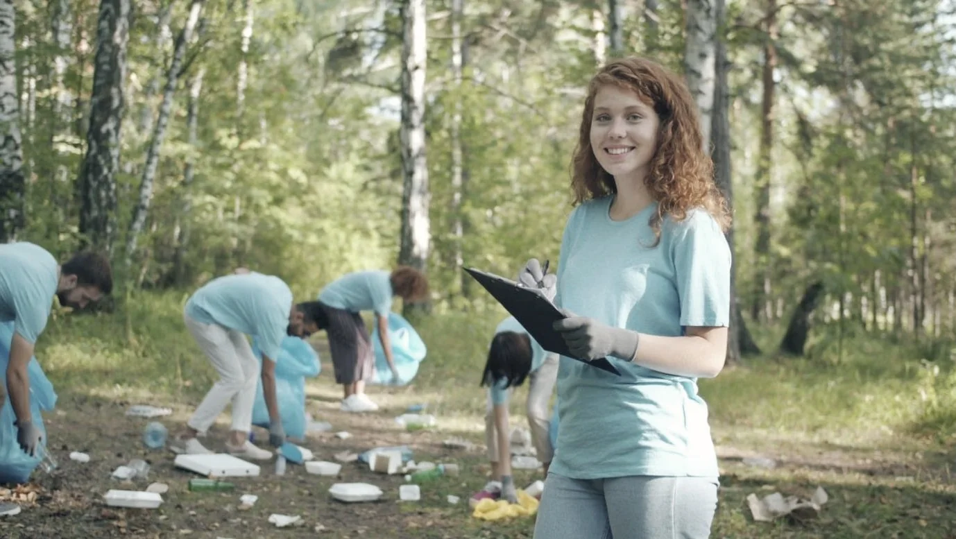 A smiling volunteer with a clipboard at a forest clean-up, with other volunteers collecting litter behind her